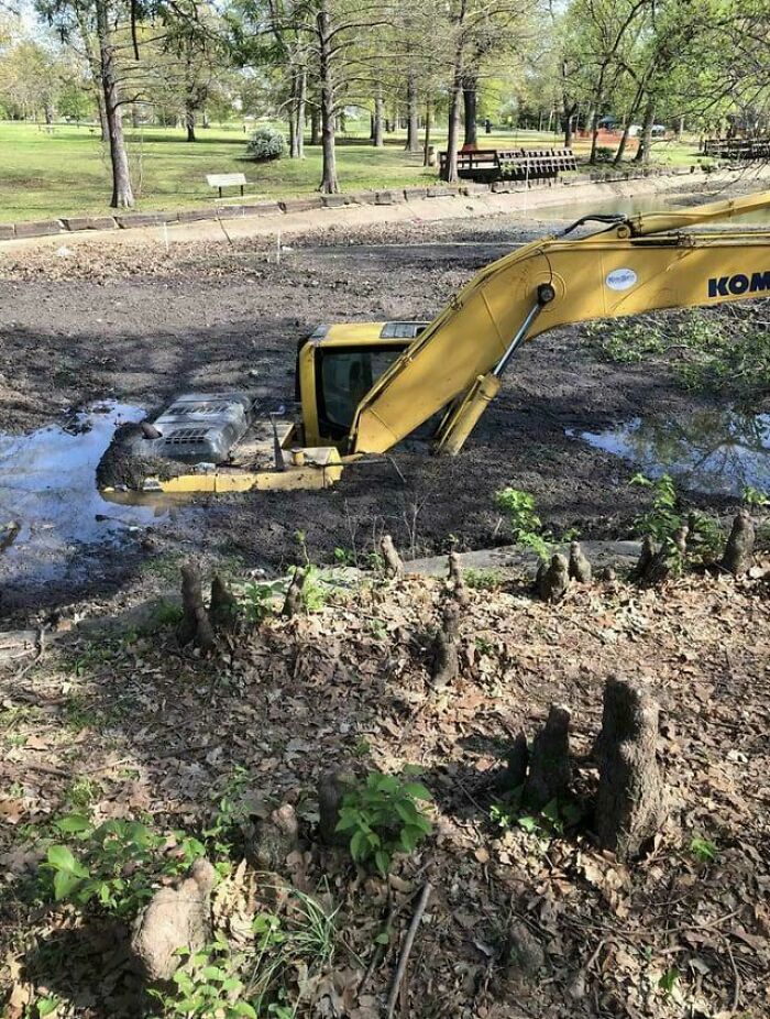 Yellow excavator stuck in muddy ground, likely an expensive incident in a park setting with trees.