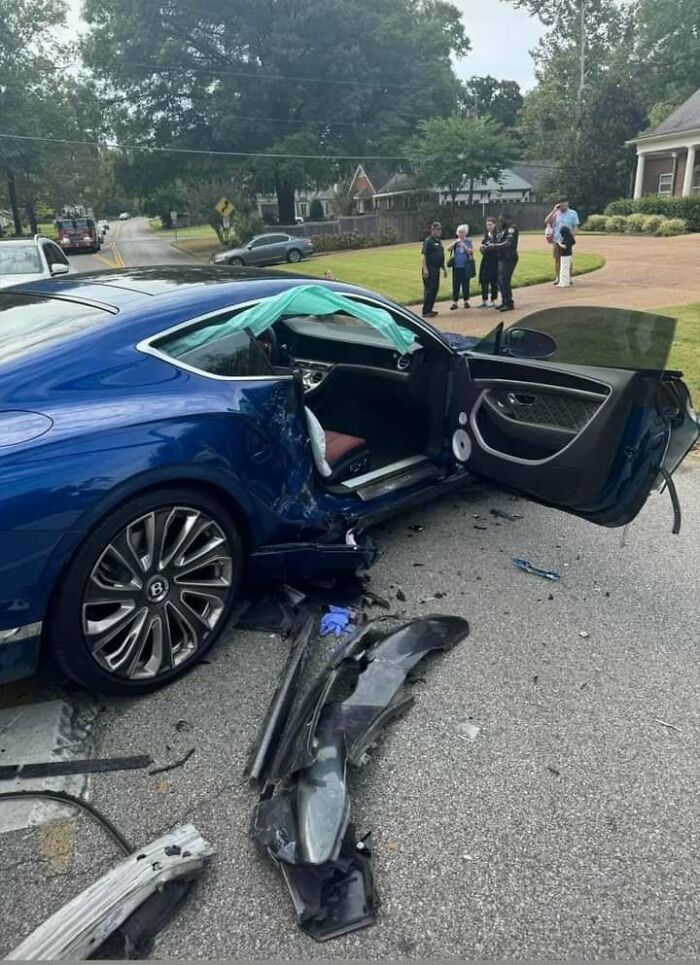 A blue luxury car badly damaged in a crash, parts scattered, likely a costly incident.