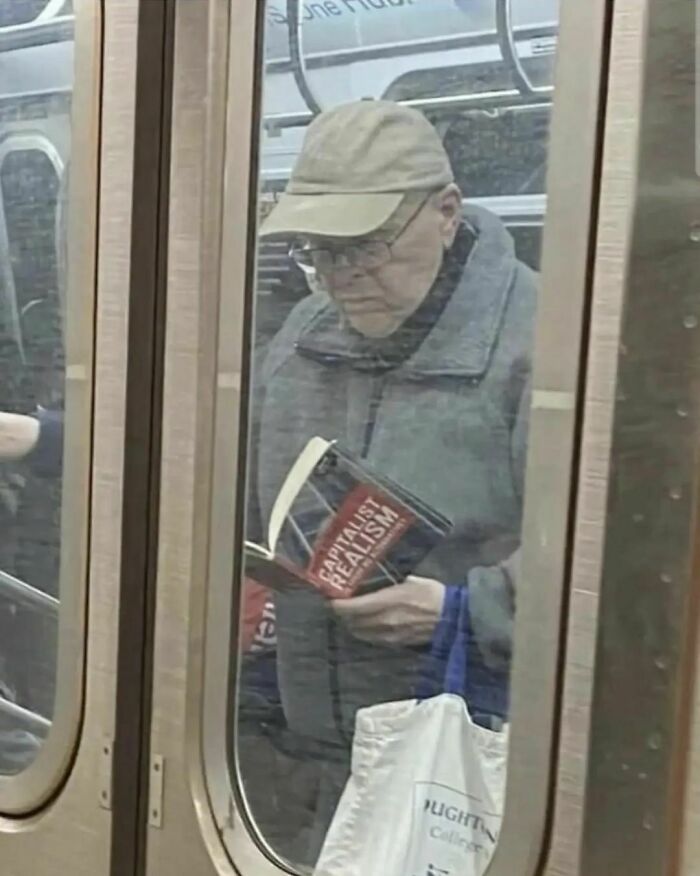 Man on subway reading "Capitalist Realism," wearing a cap and glasses, exemplifying humans in capitalism pictures.