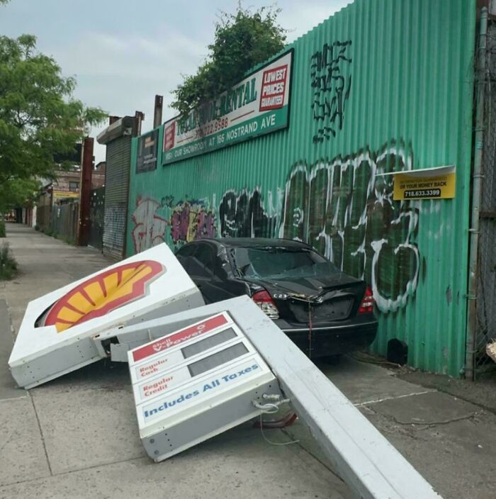 Expensive-looking incident with a fallen gas station sign damaging a car against a graffitied wall.