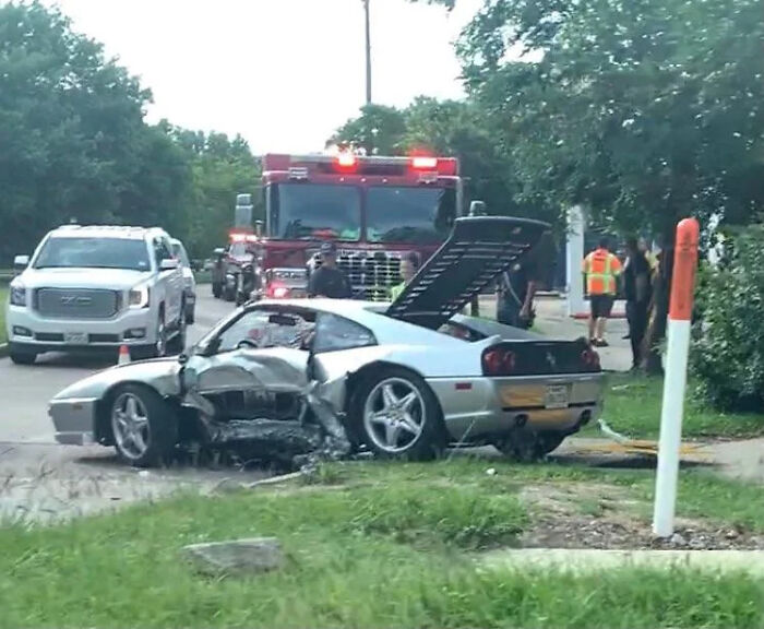 Silver sports car with severe side damage after an expensive incident, attended by emergency vehicles on a residential street.