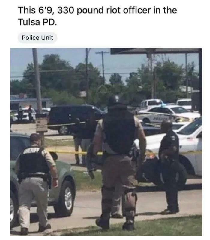 Police officer in tactical gear, showcasing absolute units things, standing among patrol cars.