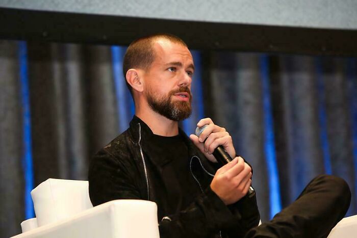 A person with a beard holding a microphone, sitting on a white chair, with a curtain backdrop at a conference.