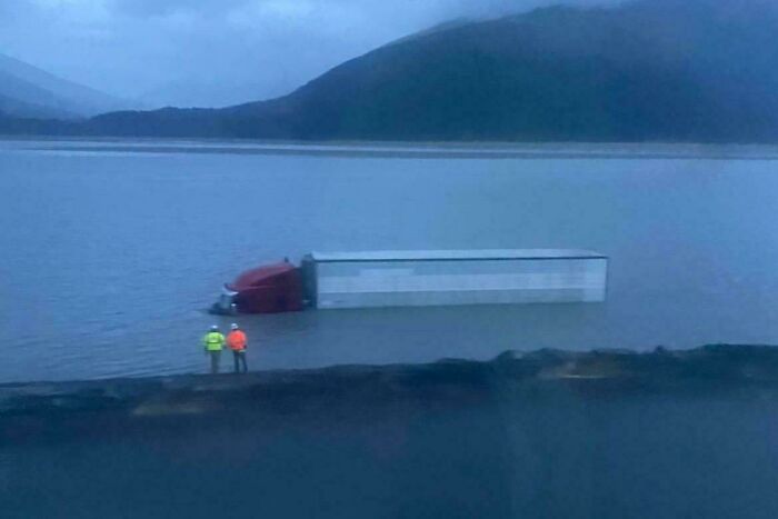 Truck submerged in water near a road, with two people in high-visibility jackets observing the incident.