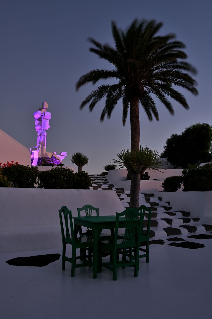 Outdoor setting with green table and chairs, palm tree, and abstract sculpture; award-winning advertising photo 2023.