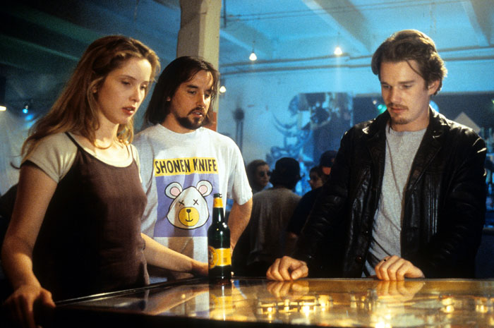 A scene from the movie Before Sunrise featuring three young adults gathered around a pinball machine. The man on the right, wearing a leather jacket, is focused on playing the game, while the woman on the left watches attentively. Another man, standing in the middle and wearing a "Shonen Knife" T-shirt, casually holds a beer bottle. The setting appears to be a lively bar or arcade with a dimly lit atmosphere.