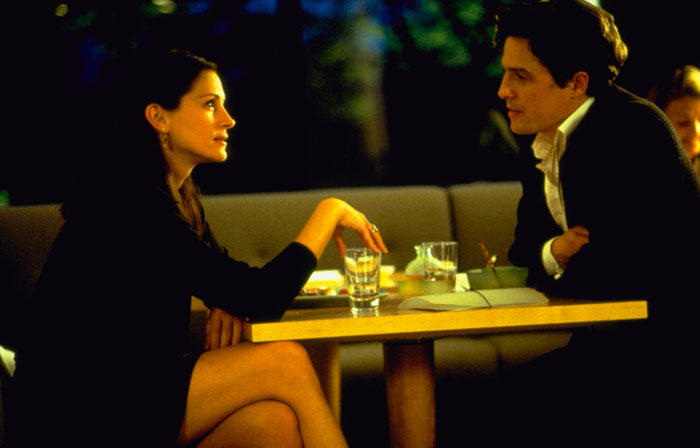 A scene from the movie Notting Hill featuring a man and a woman sitting across from each other at a small table in a dimly lit restaurant. The woman, dressed in a black outfit, leans forward slightly, engaging in conversation, while the man, wearing a suit, listens attentively with a thoughtful expression. Glasses of water and a notebook are placed on the table between them, creating an intimate and quiet atmosphere.