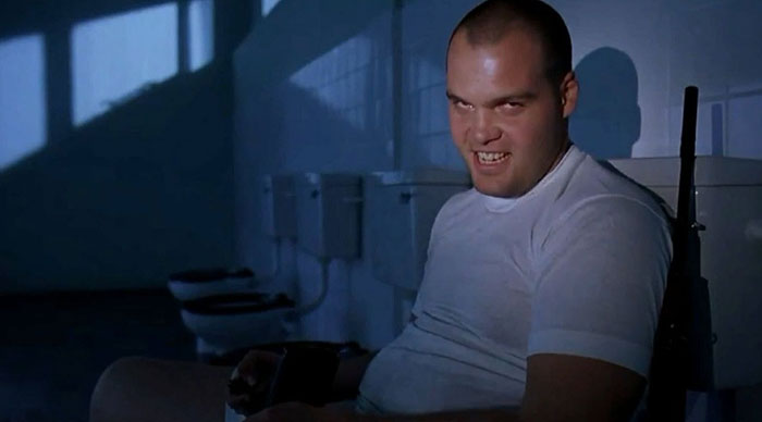  A dimly lit scene featuring a man in a white t-shirt sitting in a bathroom, with a menacing grin on his face. His intense expression, combined with the dark shadows cast in the room, creates an unsettling atmosphere. The bathroom stalls and toilets in the background add to the eerie feeling.