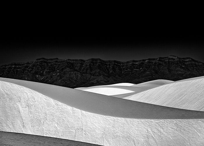 Black and white photograph of sand dunes with mountain backdrop, showcasing future photography trends, 2024 IPA winner.