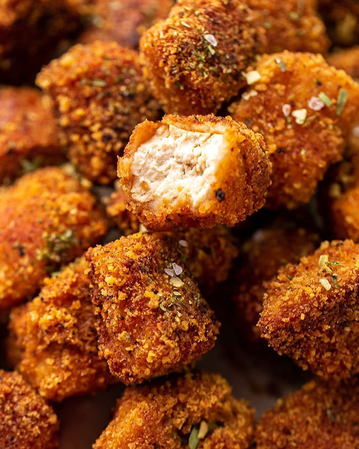 A Lunch idea featuring crispy baked tofu nuggets with a golden brown breaded coating, seasoned with herbs and spices, one nugget bitten to reveal tender tofu inside.