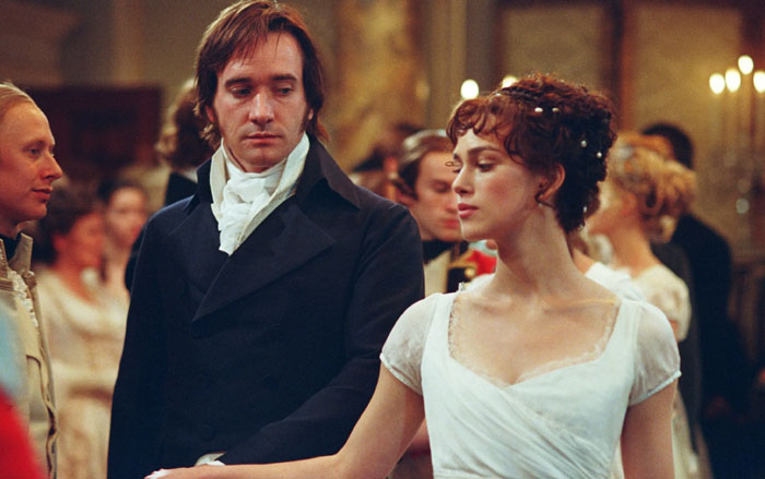 A scene from the movie Pride and Prejudice featuring a man and a woman in formal Regency-era attire at a ballroom. The man, dressed in a dark coat and white cravat, looks intently at the woman, who is wearing a white gown with her hair adorned in small flowers. She gazes away with a composed and distant expression. Other guests in period clothing can be seen in the softly lit background, enhancing the elegant, historical atmosphere of the scene.