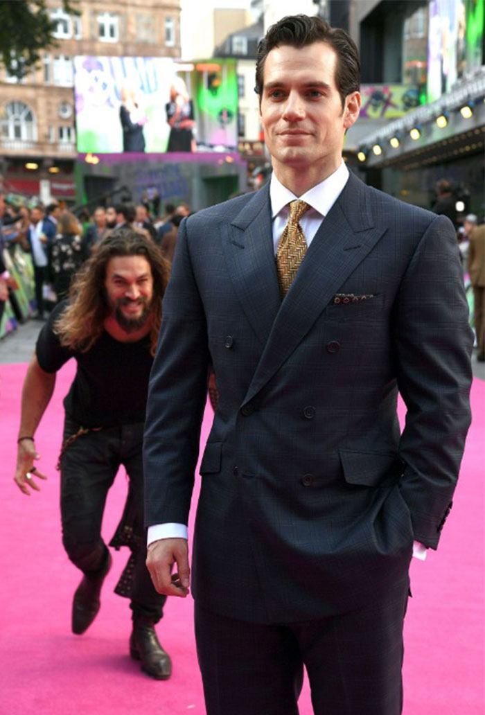 A humorous image of a well-dressed man in a suit standing confidently on a red carpet, unaware of another man playfully sneaking up behind him with a mischievous grin. The contrast between the serious demeanor of the man in the suit and the playful, stealthy approach of the man in the background creates a funny and unexpected moment. This image could inspire a "funny group chat name" related to surprise, playful antics, or unexpected photo bombs.