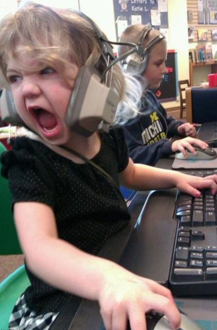 A humorous image of a young girl sitting at a computer with a large headset on, screaming with intense concentration and emotion while gripping a computer mouse. Her expression is one of fierce determination, as if she’s in the middle of a very intense gaming session or video call. Another child in the background is calmly using a computer, adding contrast to the scene. This image could serve as inspiration for a "funny group chat name" related to online gaming, passionate discussions, or chaotic energy in digital meetings.