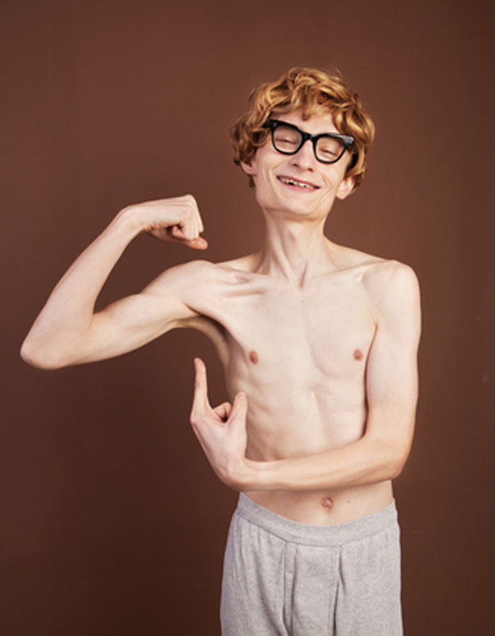 A funny image of a skinny man with glasses and a joyful smile striking a pose while flexing his bicep in a playful attempt to show off his muscles. His confident expression contrasts humorously with his thin physique. This image could be used as inspiration for a "funny group chat name" related to fitness, unexpected strength, or lighthearted self-confidence.