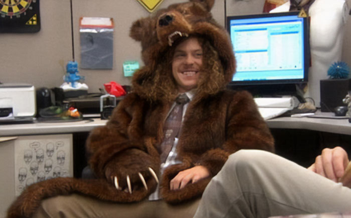 A humorous image of a man sitting in an office cubicle, smiling broadly while wearing a full bear costume with the bear's head acting as a hood. The setting is a typical office environment with computers and documents, contrasting comically with the man's unusual outfit. This image could inspire a "funny group chat name" related to office shenanigans, unique dress codes, or playful workday surprises.