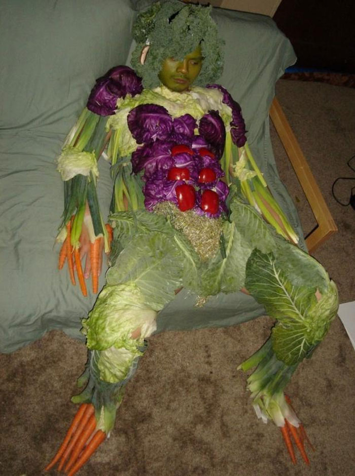 A humorous image of a person dressed entirely in vegetables, resembling a human-vegetable hybrid. Their costume includes carrots for fingers, broccoli for hair, lettuce leaves, cabbage, and red bell peppers covering the body, creating a surreal and quirky appearance. The person appears to be sitting or resting on a couch, adding to the absurdity of the situation. This image could inspire a "funny group chat name" related to vegetables, gardening, or quirky costumes.