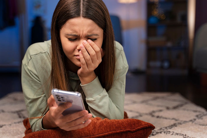 Woman lying on a rug, looking upset while reading something on her phone, hand covering part of her face.