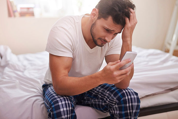 Man in a white t-shirt and plaid pants sitting on a bed, looking stressed while holding a phone, in a bedroom setting.