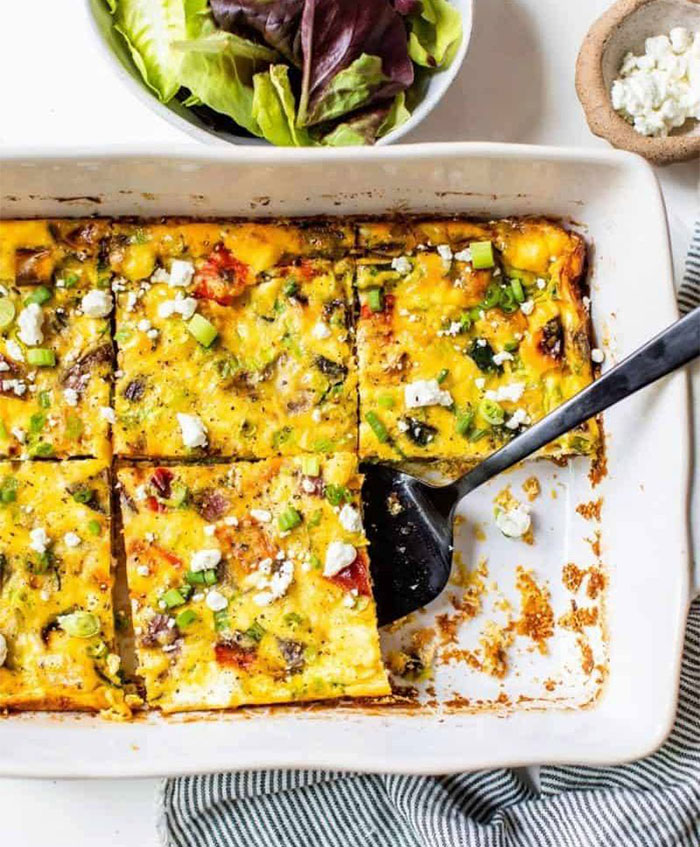 A Lunch idea featuring a colorful vegetable frittata topped with feta cheese and green onions, sliced into squares in a baking dish, with a side of fresh greens.