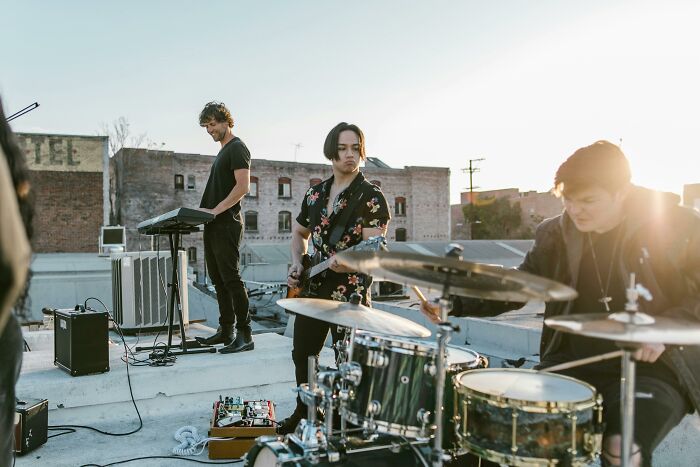 Musicians on a rooftop, with a drummer, keyboardist, and guitarist playing under the setting sun.