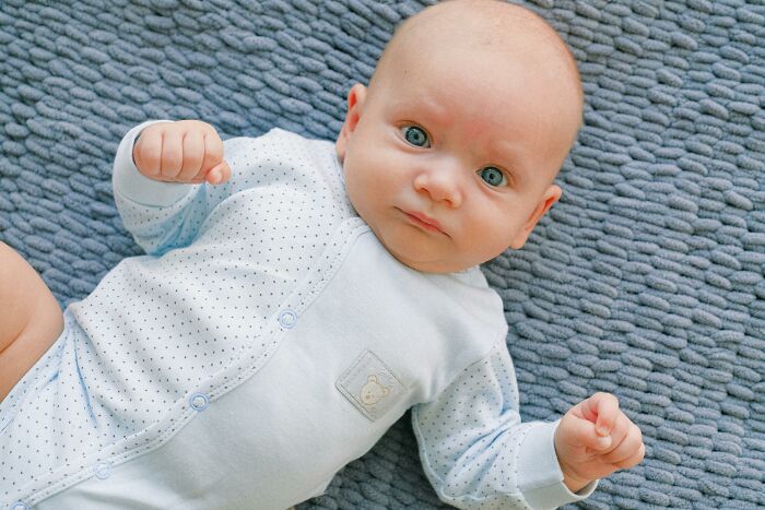 Baby lying on a gray blanket in a white onesie, giving a curious look.