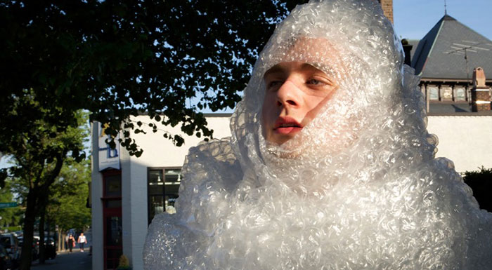  A person is fully wrapped in bubble wrap, standing outside in a sunlit street, looking off into the distance with a serious expression. The bubble wrap creates an exaggerated, protective cocoon around them, contrasting humorously with the everyday urban environment behind them. 