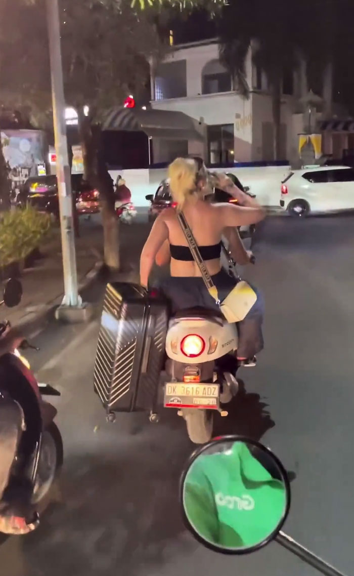 Tourist In Bali, Indonesia Filmed Riding A Scooter/Motorbike Without A Helmet, Carrying A Large Suitcase And Drinking