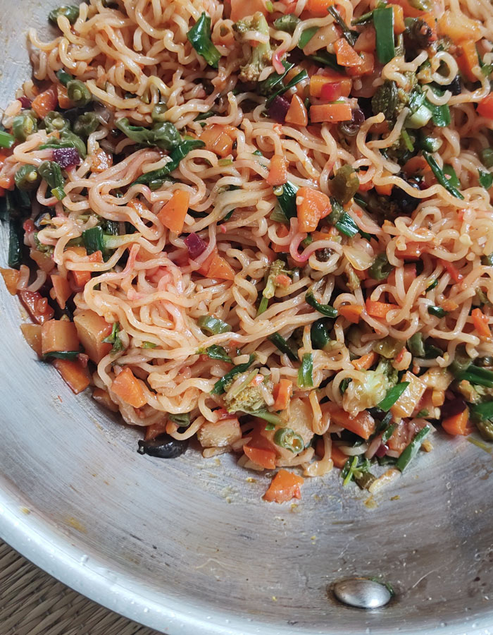 A colorful lunch idea featuring stir-fried noodles mixed with chopped vegetables, including carrots, green onions, and peas, served in a silver pan.