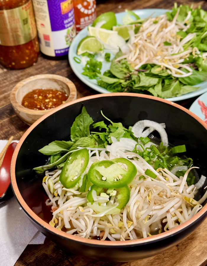 A fresh lunch idea featuring a bowl of Vietnamese pho, garnished with bean sprouts, jalapeño slices, basil, and onions. In the background, a plate with additional herbs, lime wedges, and condiments is visible alongside sauces and chili paste.