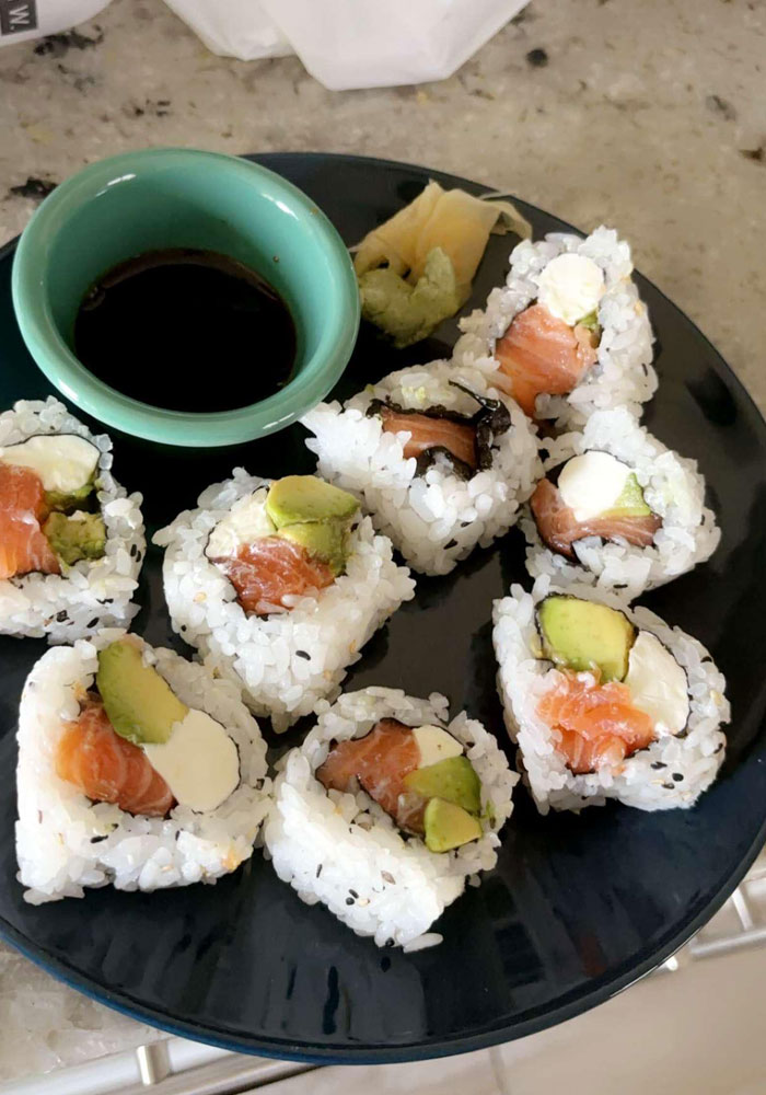 A plate of sushi rolls filled with salmon, avocado, and cream cheese, served with soy sauce, wasabi, and pickled ginger on the side. The dish is arranged on a dark plate with a small dipping bowl of soy sauce.