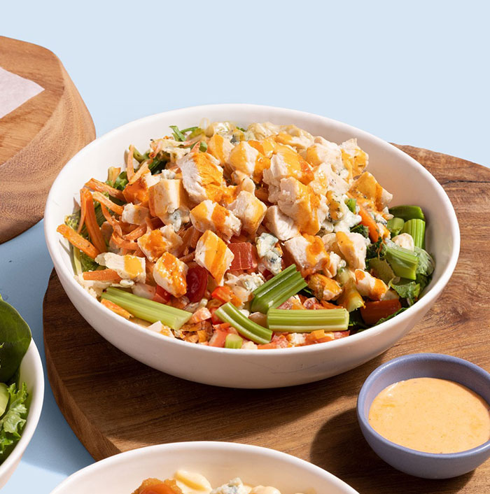 A fresh lunch idea featuring a chopped salad with grilled chicken, celery, carrots, tomatoes, and crumbled cheese, drizzled with buffalo sauce, served in a white bowl with a small side of dipping sauce.