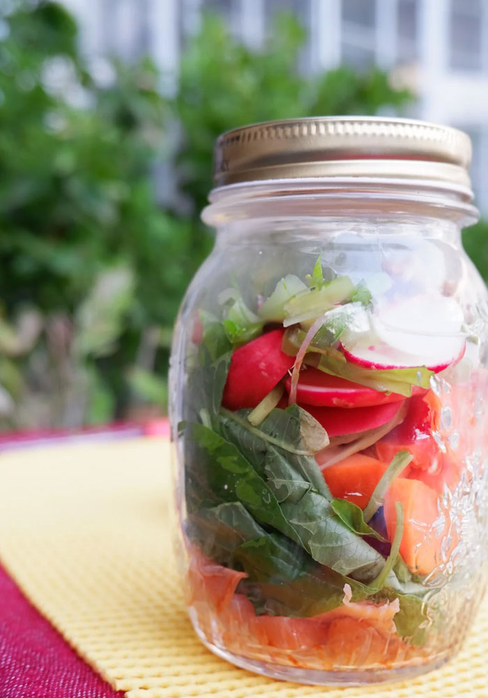 A fresh lunch idea featuring a colorful salad packed in a mason jar, with layers of leafy greens, sliced radishes, carrots, and other vegetables, placed on a yellow and red tablecloth with greenery in the background.