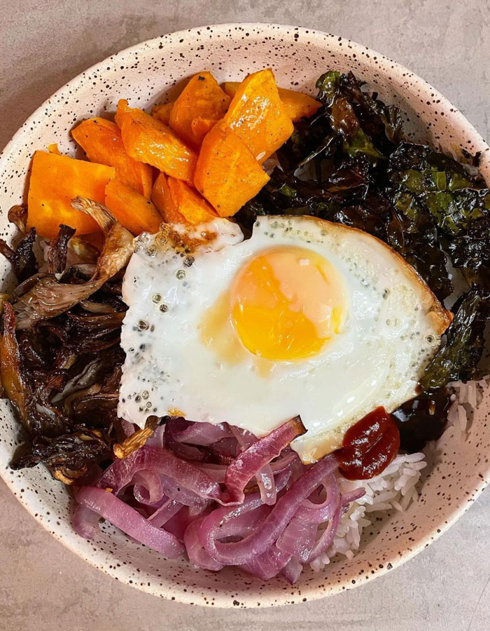 A vibrant lunch idea featuring a bowl of rice topped with a sunny-side-up egg, sautéed mushrooms, roasted sweet potatoes, pickled red onions, and a side of leafy greens.