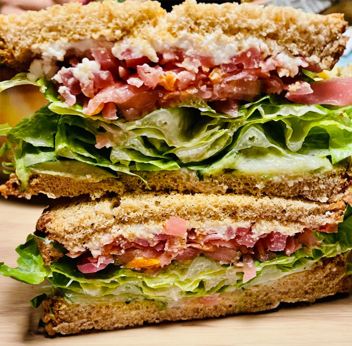 A stacked sandwich filled with layers of fresh lettuce, sliced tomatoes, cucumbers, and creamy spread, served between slices of whole wheat bread.