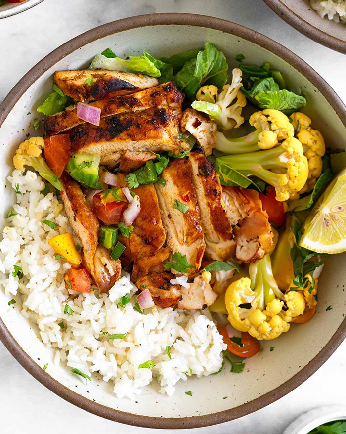 A healthy lunch idea featuring grilled chicken slices served over a bed of white rice, accompanied by roasted cauliflower, fresh lettuce, diced vegetables, and a lemon wedge, all arranged in a bowl.