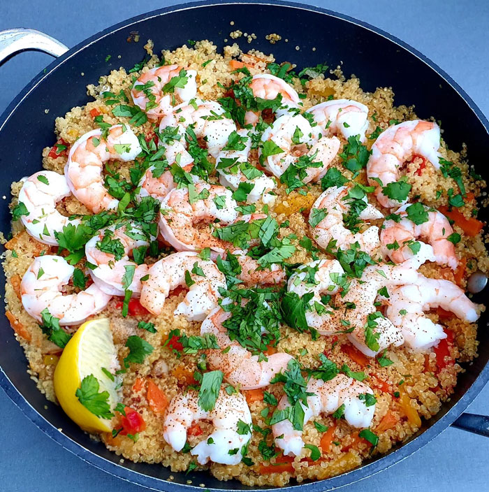 A healthy lunch idea featuring a pan of cooked quinoa topped with seasoned shrimp, garnished with fresh parsley and served with a lemon wedge.