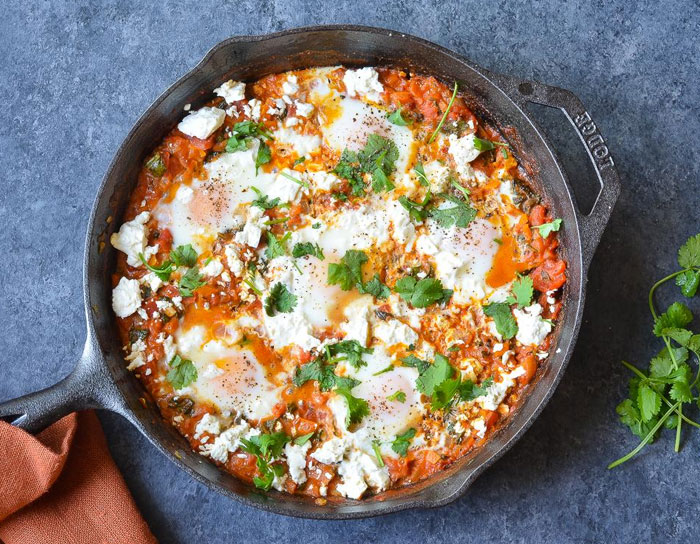 A hearty lunch idea featuring shakshuka in a cast iron skillet, with poached eggs nestled in a spiced tomato sauce, topped with crumbled cheese and fresh cilantro.