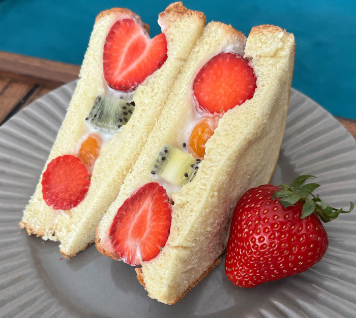 A light and refreshing lunch idea featuring two fruit sandwiches filled with fresh strawberries, kiwi, and cream, served on soft white bread, with a whole strawberry on the side, placed on a gray plate.