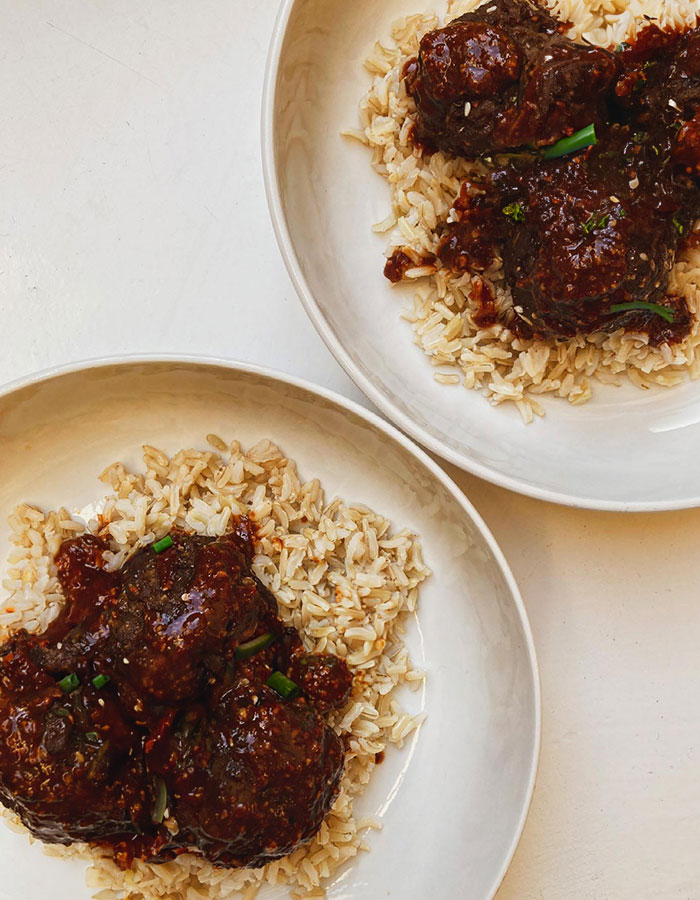 A hearty lunch idea featuring two bowls of brown rice topped with tender meatballs coated in a rich, dark sauce, garnished with fresh herbs, served in white bowls.