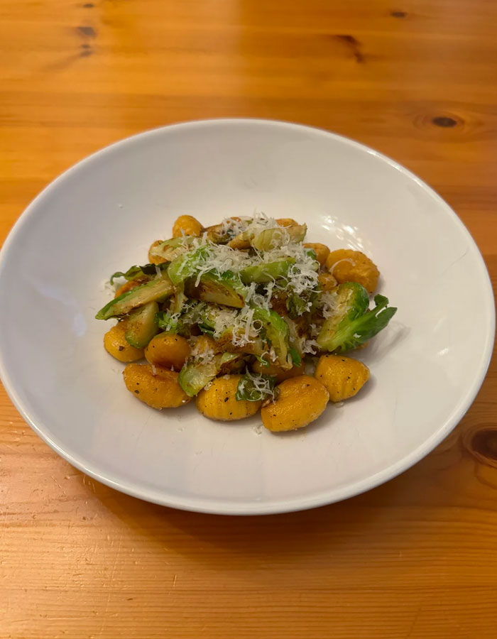 A gourmet lunch idea featuring a plate of pan-fried gnocchi with sautéed Brussels sprouts, topped with grated Parmesan cheese, served in a white bowl on a wooden table.