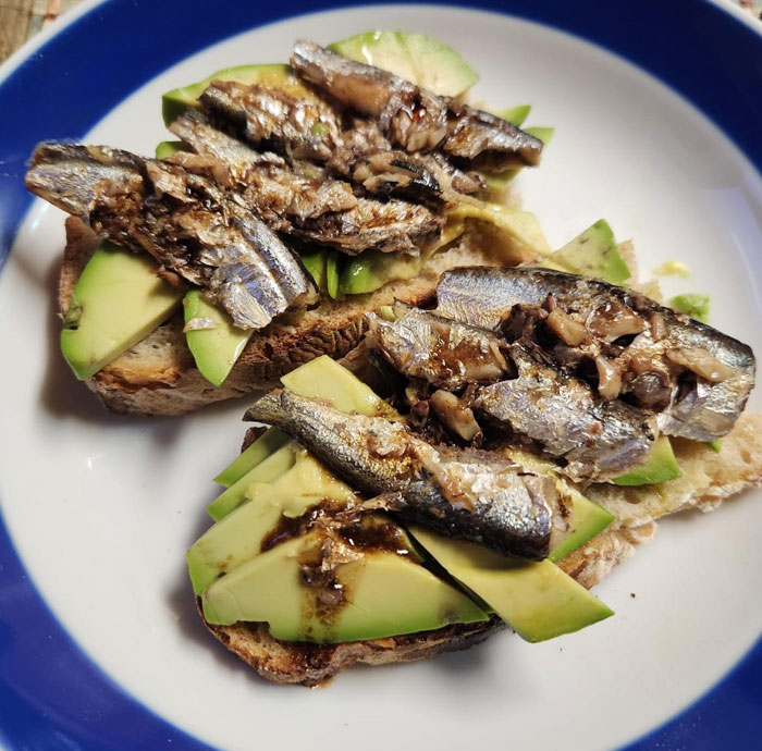 A simple lunch idea featuring toasted bread topped with slices of avocado and sardines, drizzled with a light sauce, served on a white and blue plate.