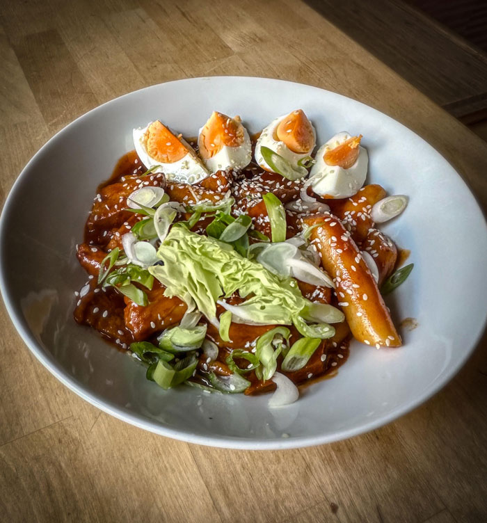 Lunch idea featuring a bowl of tteokbokki topped with sesame seeds, sliced green onions, cabbage, and halved boiled eggs, served on a wooden table.