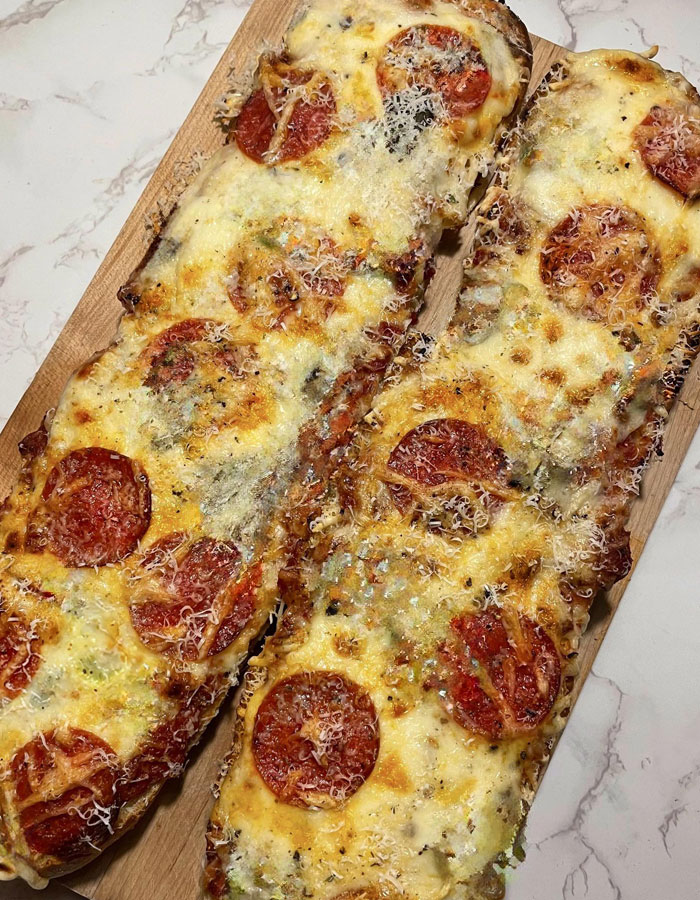 Lunch idea featuring two long flatbread garlic bread pizzas topped with pepperoni, melted cheese, and grated Parmesan on a wooden serving board placed on a marble surface.