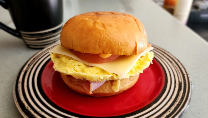 A Lunch idea featuring a breakfast sandwich with scrambled eggs, ham, cheese, and tomato on a toasted bun, served on a red and black plate.