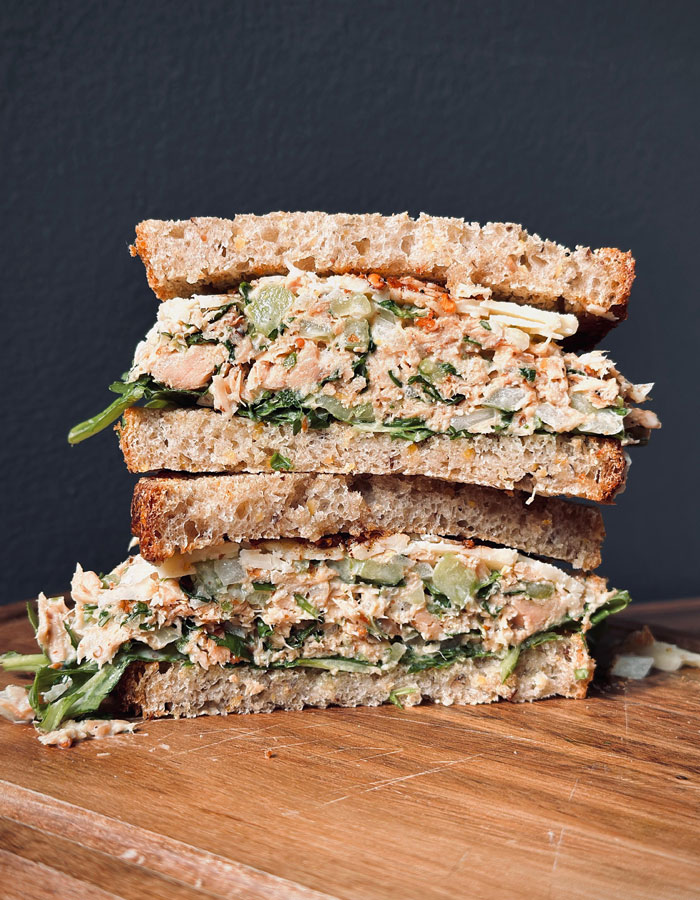 A Lunch idea featuring a hearty sandwich with tuna salad, greens, and vegetables stacked between two slices of whole grain bread on a wooden surface.