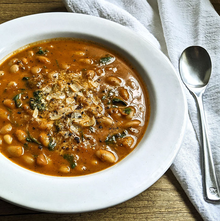 A lunch idea featuring a hearty bowl of bean soup with a rich tomato-based broth, topped with grated cheese and pepper, served with a spoon on the side.