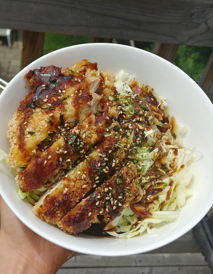 A flavorful lunch idea featuring crispy breaded chicken cutlets drizzled with sauce, served on a bed of shredded cabbage, and garnished with sesame seeds and herbs in a white bowl.