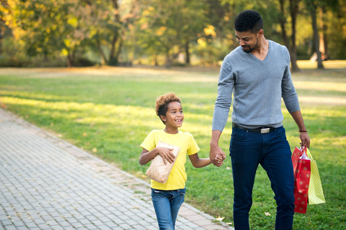 Father and son walking in a park, enjoying a sunny day together, carrying shopping bags and snacks.