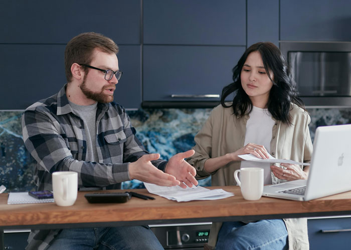 Couple discussing financial decisions at a wooden table with coffee mugs and a laptop in a modern kitchen.