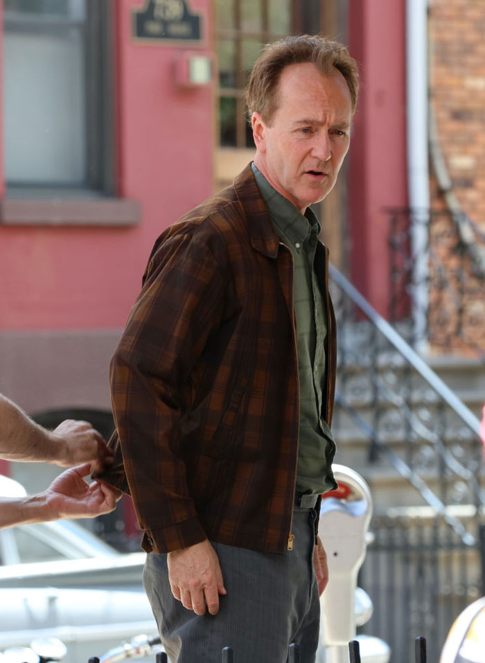  A B list celebrity, Edward Norton, wearing a plaid brown jacket and a green shirt is standing on a city sidewalk, with a person adjusting his clothing.